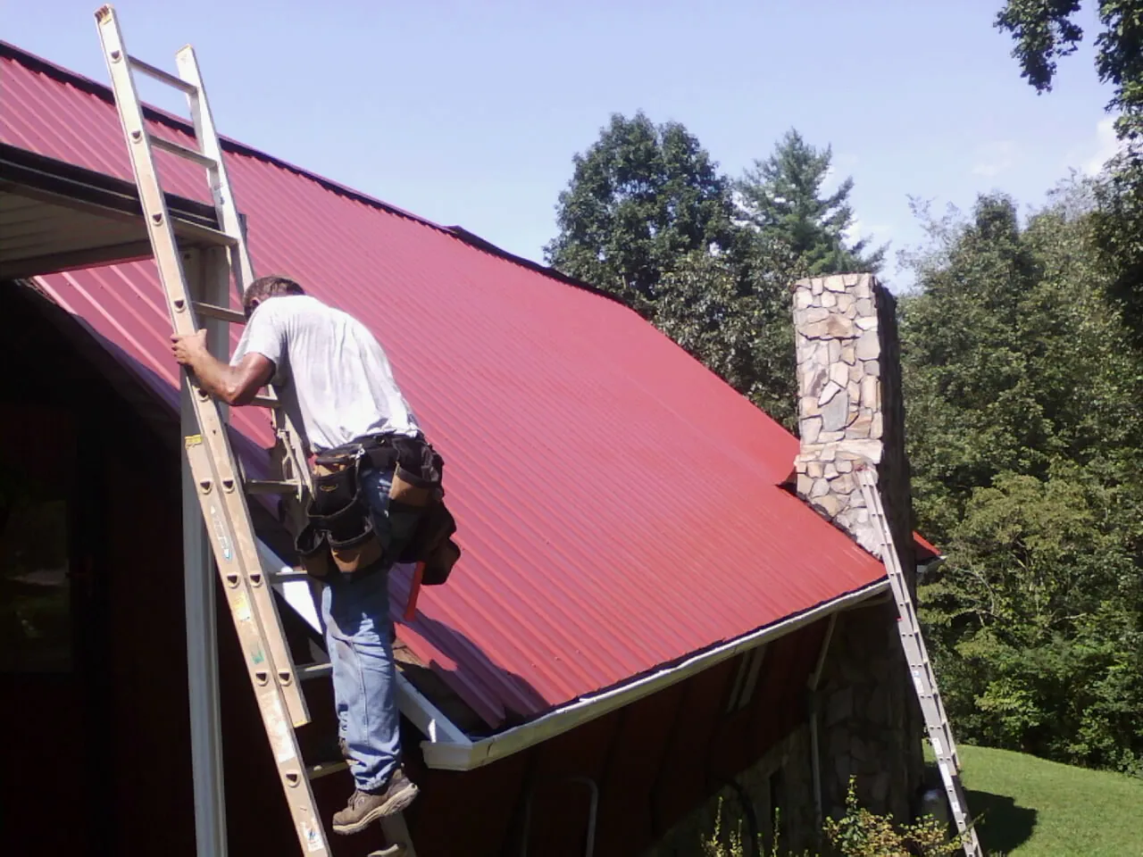 Roofer on a ladder beside a red metal roof and stone chimney — Soffit & Fascia Repair by Roofing Brothers in Pocomoke City